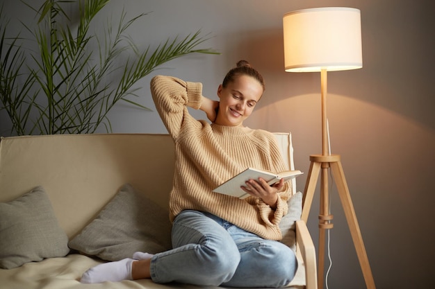 Foto einer entzückten, entspannten Frau mit beigefarbenem Pullover und Jeans, die auf Husten sitzt und entspannt ein Buch liest oder studiert und in die Kamera blickt und dabei einen positiven Ausdruck ausdrückt