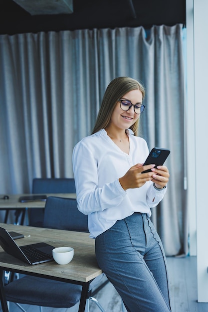 Foto einer erfolgreichen jungen Frau in einem Hemd, die lächelt und an einem Laptop arbeitet, während sie in einem modernen Büro mit großen Fenstern telefoniert Fernarbeit mit Kaffee