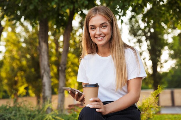 Foto einer erfreuten positiven optimistischen jungen Dame sitzt im Naturpark mit trinkendem Kaffee des Handys.