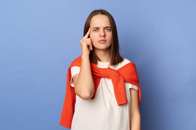 Foto einer ernsthaft konzentrierten jungen erwachsenen Frau, die ein weißes T-Shirt und einen orangefarbenen Pullover über den Schultern trägt und auf ihre Schläfe zeigt und in die Kamera blickt, die isoliert auf blauem Hintergrund posiert