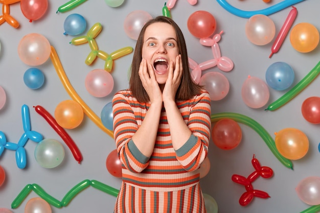 Foto einer extrem fröhlichen Frau mit braunen Haaren, die ein gestreiftes Kleid trägt, die um bunte, aufgeblasene Luftballons herumsteht und vor Glück schreit und erstaunt ist, wie sie ihr Gesicht berührt