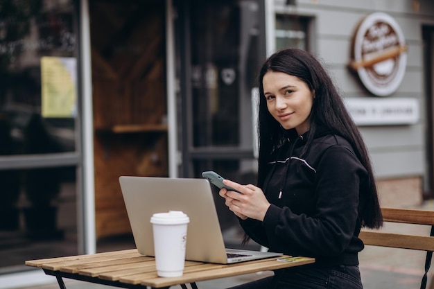 Foto einer freiberuflichen Frau, die Kaffee zum Mitnehmen genießt, während sie im Freien an einem tragbaren Computer arbeitet, der mit öffentlichem WLAN verbunden ist Stilvolle Studentin, die online studiert, während sie Freizeit in einem Park verbringt