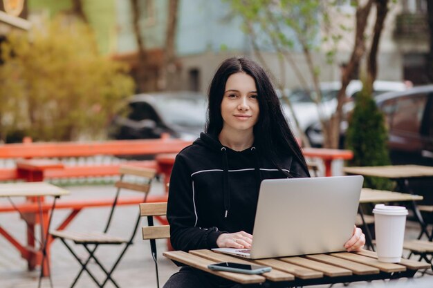 Foto einer freiberuflichen Frau, die Kaffee zum Mitnehmen genießt, während sie im Freien an einem tragbaren Computer arbeitet, der mit öffentlichem WLAN verbunden ist Stilvolles Studentenmädchen, das online studiert, während es Freizeit in einem Park verbringt