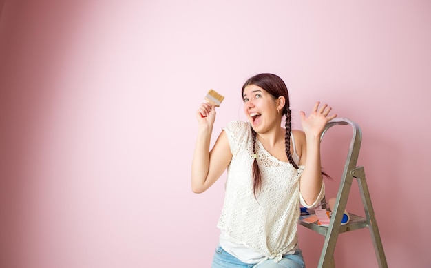 Foto einer fröhlichen Frau mit Bürste neben Trittleiter und Rolle vor leerer rosa Wand