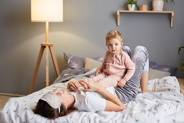 Foto einer glücklichen Familienmutter mit ihrer kleinen Tochter, die im Schlafzimmer spielt, während sie auf dem Bett liegt, ein Kind, das auf Mama sitzt und mit ernster Miene wegschaut