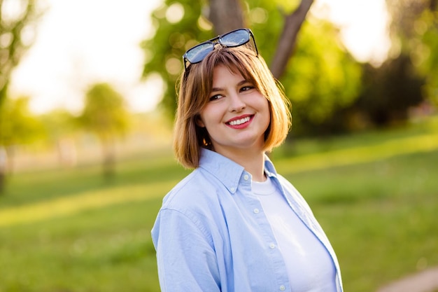 Foto einer hübschen, glänzenden jungen Frau, die ein blaues Hemd trägt und lächelt, während sie außerhalb des grünen Parks spazieren geht