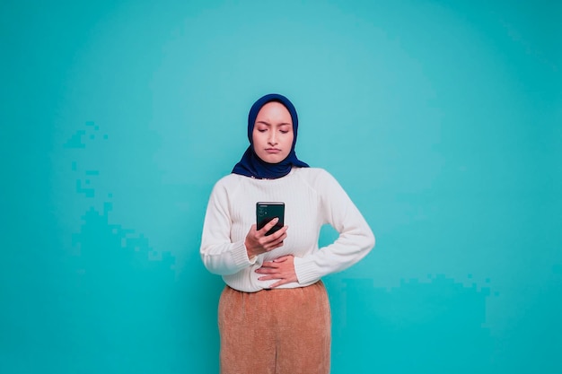 Foto einer hungrigen aufgeregten jungen muslimischen Frau, die ihr Telefon hält und sich fragt, was sie leckeres Essen bestellen soll, isoliert auf blauem Hintergrund