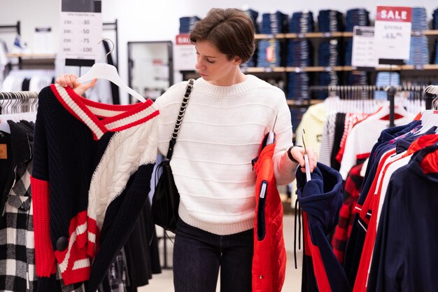 Foto einer jungen attraktiven brünetten Frau mit kurzem Haarschnitt in einem weißen Pullover wählt stilvolle und lässige Kleidung in einem Geschäft in einem Einkaufszentrum. Shop-Interieur