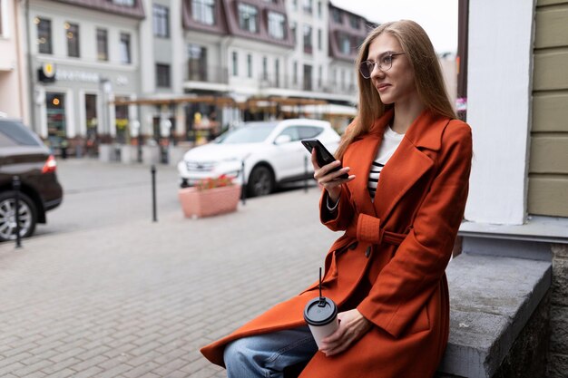 Foto einer jungen Bloggerin, die mit einem Telefon in der Hand auf der Veranda eines Gebäudes sitzt
