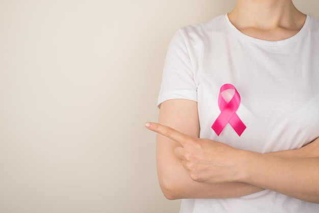 Foto einer jungen Frau in weißem T-Shirt mit rosa Schleifensymbol für Brustkrebsbewusstsein, die mit dem Zeigefinger auf isoliertem grauem Hintergrund mit Leerzeichen auf etwas zeigt