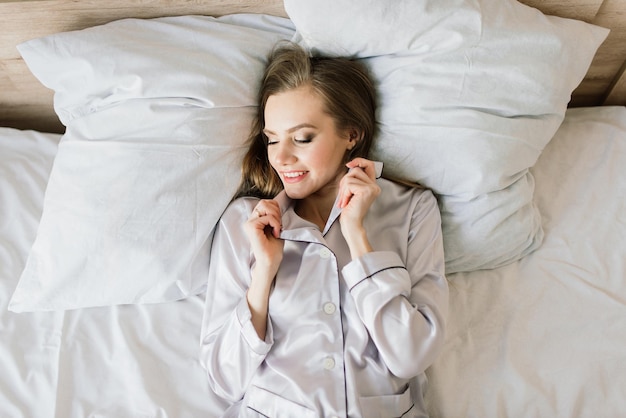 Foto einer jungen fröhlichen Frau im Pyjama mit Laptop und lächelnd beim Sitzen auf dem Bett im hellen Raum