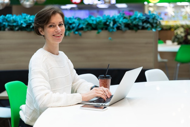 Foto einer jungen glücklichen, positiv lächelnden Frau, die im Food Court eines Einkaufszentrums an einem Tisch sitzt und an einem Computer-Laptop arbeitet. Freiberufliches Konzept