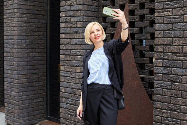 Foto einer jungen, positiven, fröhlichen, zufriedenen Frau, die im Freien auf der Straße spaziert, ein Selfie mit der Kamera macht Schöne blonde Frau, die im Business-Stil durch die Straßen der Stadt geht