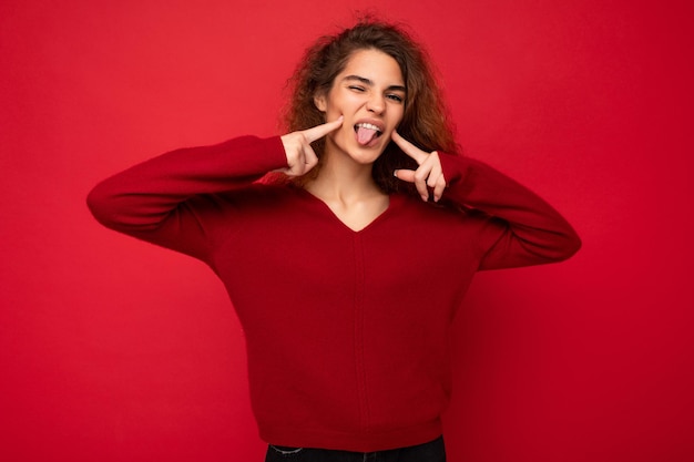 Foto einer jungen, positiven, glücklichen, schönen Brünetten, lockige Frau mit aufrichtigen Emotionen, die einen lässigen roten Pullover trägt, der auf rotem Hintergrund mit Kopienraum isoliert ist und Tingue zeigt.