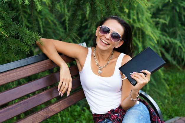 Foto einer jungen schönen Frau, die auf einer Holzbank im Park sitzt, mit schwarzem Notizblock in der Hand in Cas...