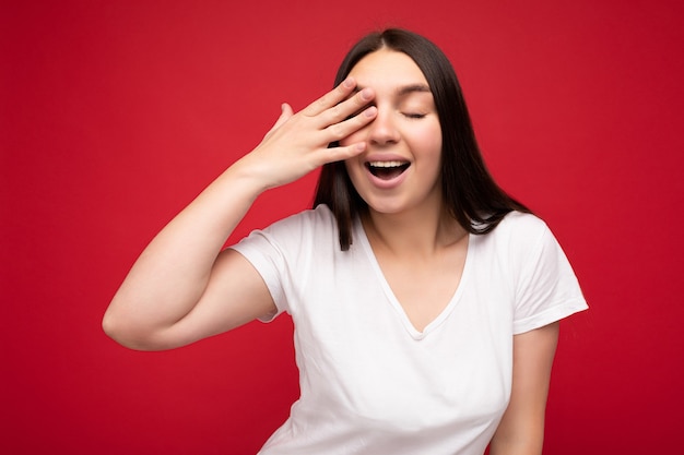 Foto einer positiven jungen, schönen brünetten weiblichen Person mit aufrichtigen Emotionen, die ein lässiges weißes T-Shirt für das Modell trägt, einzeln auf rotem Hintergrund mit Kopienraum und Augen bedeckend.