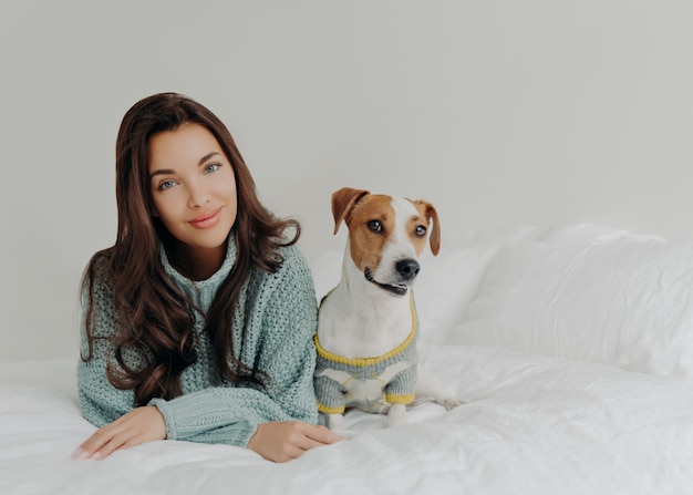 Foto einer schönen dunkelhaarigen Frau kleidet ihren Hund für kaltes Wetter an, geht im Freien spazieren, liegt zusammen am bequemen Bett