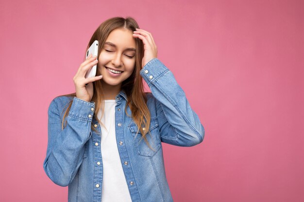 Foto einer schönen glücklich lächelnden jungen blonden Frau, die ein lässiges blaues Jeanshemd trägt, isoliert auf rosa Hintergrund, das in der Hand hält und mit geschlossenen Augen auf dem Handy spricht