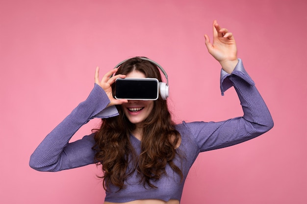 Foto einer schönen, glücklichen, jungen, brünetten, lockigen Frau, die ein lila Crop-Top trägt, isoliert über einer rosafarbenen Hintergrundwand, die ein Mobiltelefon mit leerem Display und Bluetooth-Kopfhörern hält und vorschlägt