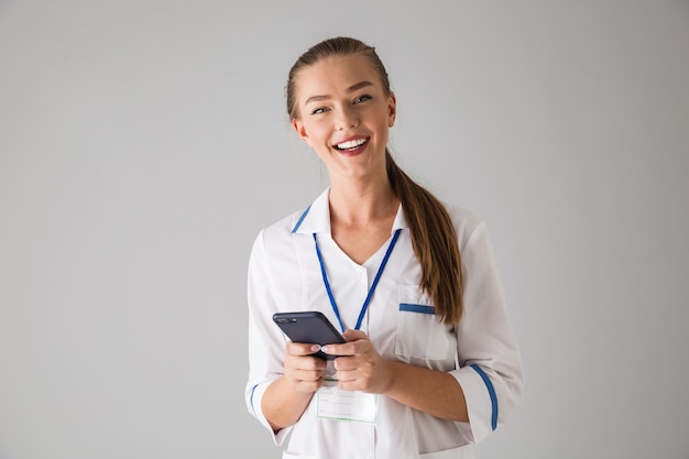 Foto einer schönen glücklichen jungen Frau Kosmetikerin Arzt isoliert über graue Wand hält Handy mit.