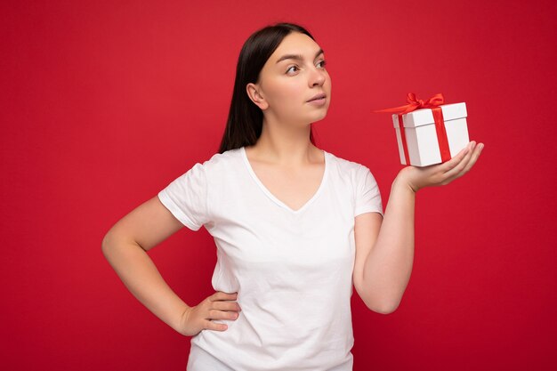 Foto einer schönen jungen brünetten Frau, die über einer roten Hintergrundwand isoliert ist und ein weißes, lässiges T-Shirt für ein Modell trägt, das eine weiße Geschenkbox mit rotem Band hält und zur Seite schaut