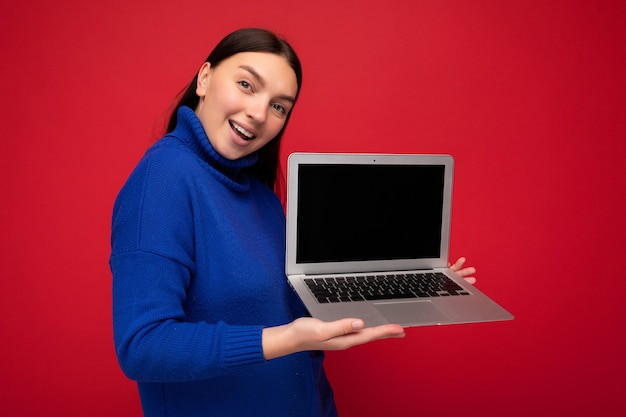 Foto einer schönen jungen Frau, die einen Computer-Laptop hält und in die Kamera schaut, die über buntem Hintergrund isoliert ist?