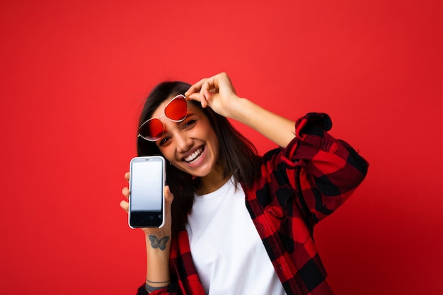 Foto einer schönen lächelnden, glücklichen jungen Frau, die gut aussieht und ein lässiges, stilvolles rotes Hemd, weißes T-Shirt und eine rote Sonnenbrille trägt, die isoliert auf rotem Hintergrund mit Kopienraum mit Smartphone-Show steht