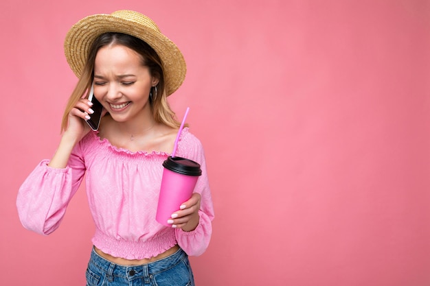 Foto einer schönen positiven emotionalen jungen blonden Frau mit rosafarbener Bluse und Strohhut, die auf dem Handy spricht, während sie Getränke trinkt, einzeln auf rosafarbenem Hintergrund mit Kopienraum.