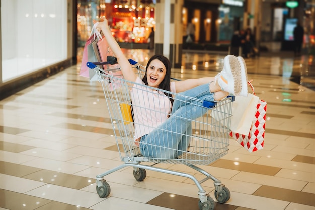 Foto einer süßen, aufgeregten Frau, die in einem Einkaufswagen sitzt und in einem Supermarkt lächelt