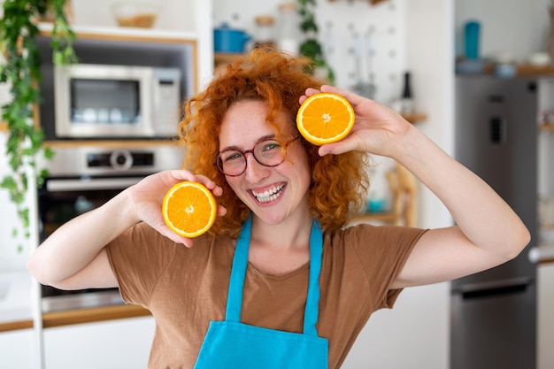 Foto einer süßen kaukasischen Frau, die lächelt und zwei orangefarbene Teile hält, während sie zu Hause Gemüsesalat in der Küche kocht