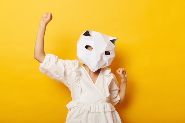 Foto einer überglücklichen anonymen Frau, die Kleid und Papierkatzenmaske trägt, hob ihre Fäuste und feierte ihren Erfolg und Sieg, während sie isoliert vor gelbem Hintergrund posierte
