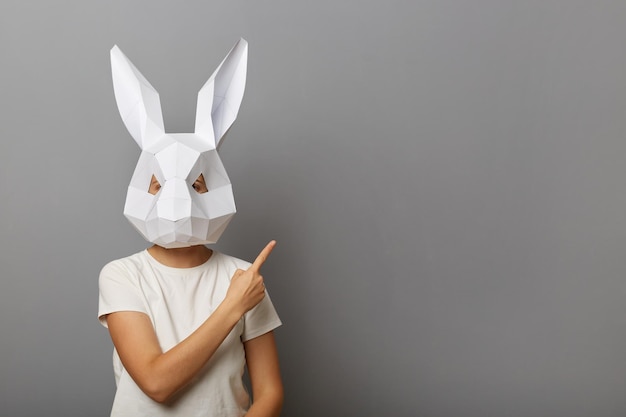 Foto einer unbekannten Frau mit weißem T-Shirt und Hasenmaske aus Papier, die mit dem Zeigefinger auf den Werbebereich zeigt, Kopienraum für Werbezwecke, der isoliert vor grauem Hintergrund steht