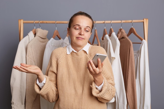 Foto einer unsicheren, verwirrten Frau, die mit Outfits vor einem Kleiderbügel steht und mit dem Smartphone die Schultern zuckt, weiß keine Antwort beim Einkaufen in einem Modegeschäft