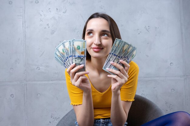 Foto einer wohlhabenden Frau in einfacher Kleidung, die einen Fan von Dollargeld hält, isoliert vor einem Betonwandhintergrund