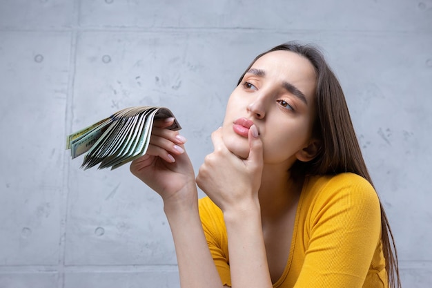 Foto einer wohlhabenden Frau in einfacher Kleidung, die einen Fan von Dollargeld hält, isoliert vor einem Betonwandhintergrund