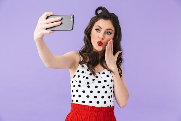 Foto einer wunderschönen Pin-up-Frau in einem Vintage-Polka-Dot-Kleid, das ein Selfie-Foto auf einem schwarzen Smartphone hält und über der violetten Wand isoliert