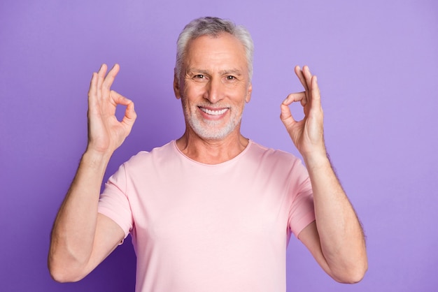 Foto eines alten Mannes im Ruhestand, der zwei Hände hebt, zeigt ein weißes, lächelndes, rosa T-Shirt mit isoliertem violetten Farbhintergrund