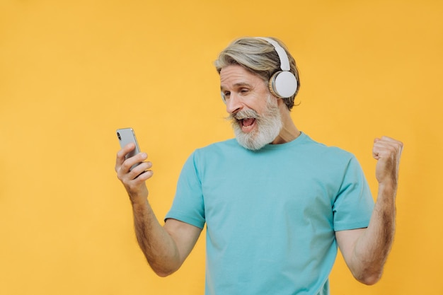Foto eines ausdrucksstarken grauhaarigen Senioren mit Kopfhörern und einem Telefon in den Händen isoliert auf gelbem Hintergrund