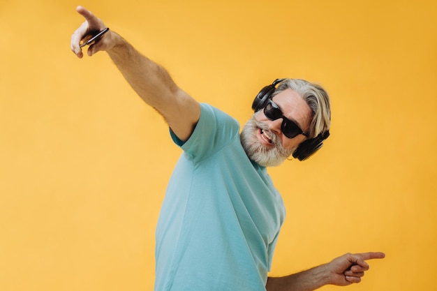 Foto eines ausdrucksstarken grauhaarigen Senioren mit Kopfhörern und einem Telefon in den Händen isoliert auf gelbem Hintergrund