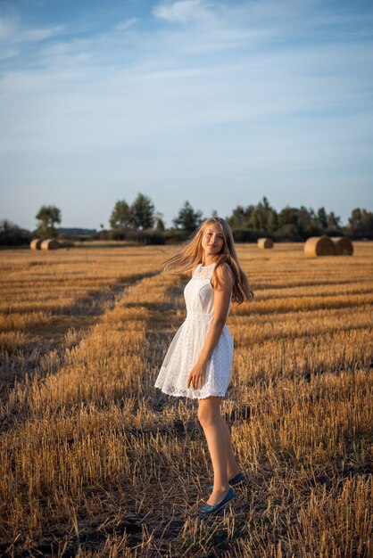 Foto eines blonden Mädchens, das ein weißes Kleid trägt, das im Feld des geernteten Roggens geht