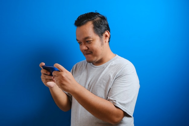 Foto eines dicken asiatischen Mannes, der ein graues T-Shirt trägt und glücklich aussieht, wenn er ein Spiel auf seinem Smartphone spielt. Männer zeigen selbstbewusste Gesten. isoliert auf blauem Hintergrund