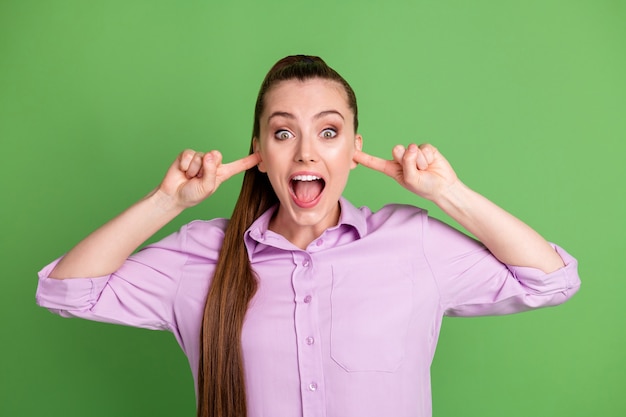 Foto eines erstaunten Mädchens, das die Ohren mit dem Zeigefinger schreit, beeindruckt, trägt ein violettes Outfit, das über grünem Hintergrund isoliert ist