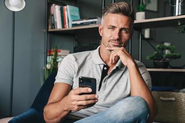 Foto eines erwachsenen kaukasischen Mannes in Freizeitkleidung, der ein Smartphone hält, während er zu Hause auf einem Sessel sitzt