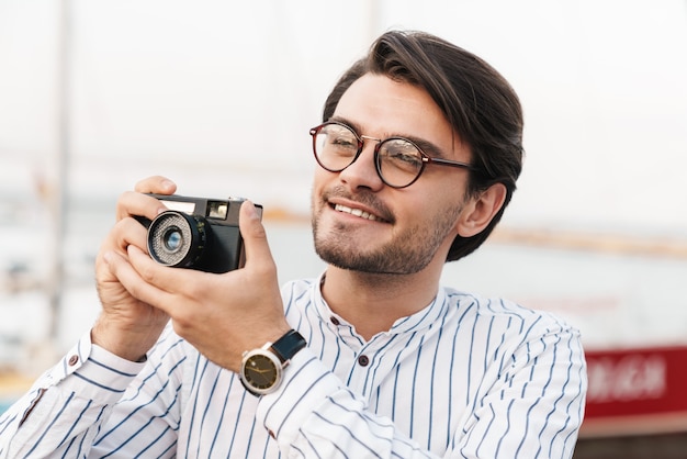 Foto eines fröhlichen kaukasischen Mannes mit Brille, der ein Foto mit einer Retro-Kamera macht und beim Gehen auf dem Pier lächelt