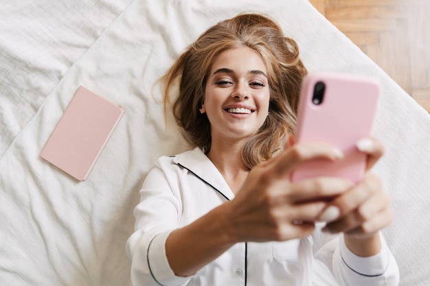Foto eines glücklich lächelnden jungen süßen Mädchens im Pyjama zu Hause auf dem Bett mit Handy.