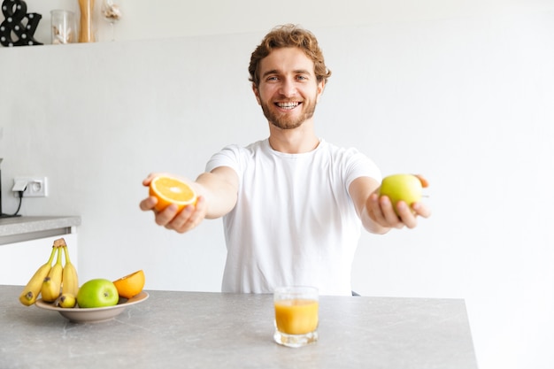 Foto eines glücklichen jungen bärtigen Mannes am Tisch zu Hause, der Früchte hält.