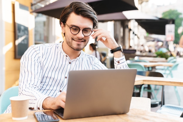 Foto eines glücklichen kaukasischen Mannes mit Brille, der auf dem Laptop tippt und das Handy benutzt, während er im Café draußen Kaffee trinkt
