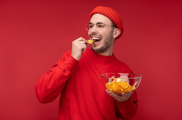 Foto eines glücklichen Mannes mit Bart in Brille und roter Kleidung. Hält und isst einen Teller mit Chips, auf rotem Hintergrund isoliert.