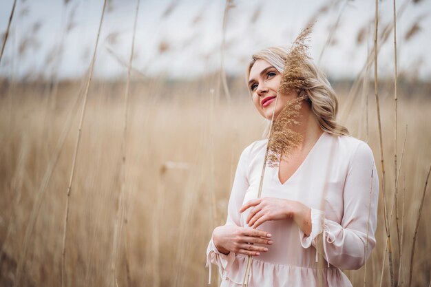 Foto eines hübschen lächelnden Mädchens mit langen blonden lockigen Haaren in hellen langen Drees, die in einem Schilffeld stehen