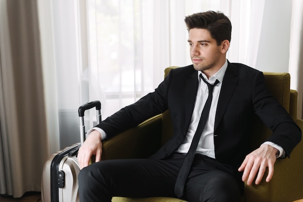 Foto eines jungen beruhigenden Geschäftsmannes im schwarzen Anzug, der beiseite schaut und auf einem Sessel mit Koffer in der Hotelwohnung sitzt?